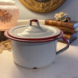 Op de afbeelding: Witte emaille kamerpot met deksel en handvat. De pot heeft een rode rand rond de rand en op het handvat. De achtergrond omvat een vintage lamp en boeken.