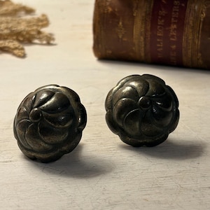 Peut inclure: Deux boutons de meuble antiques de couleur bronze avec un motif floral. Les boutons sont ronds et présentent une surface texturée et détaillée. L'arrière-plan comprend une pile de vieux livres avec des lettres dorées.