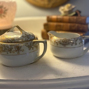 May include: A white porcelain sugar bowl with a lid and a creamer. Both pieces are decorated with gold filigree and trim. The sugar bowl has a handle on the lid and the creamer has a handle on the side.