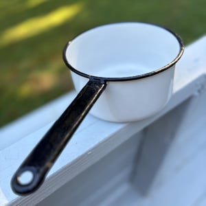 Könnte beinhalten: Ein weißer Emaille-Kochtopf mit schwarzem Griff und Rand. Der kleine Kochtopf steht auf einer weißen Oberfläche, mit einem verschwommenen grünen Hintergrund. Der Griff hat ein Loch am Ende zum Aufhängen.