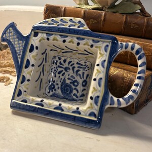 May include: A ceramic watering can shaped dish featuring a hand-painted blue and yellow floral pattern. The square-shaped basin has a central blue floral motif, a curved handle on the right, and a small spout on the left with a blue grid design.