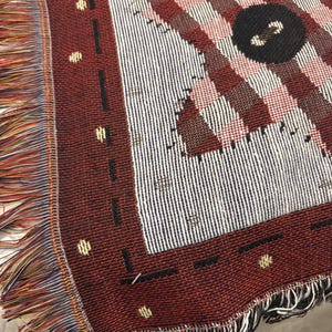 May include: A woven throw blanket with a geometric pattern in shades of red, white, and black. The blanket features a fringed edge with multiple colors. The design includes stripes, squares, and a large black circle.