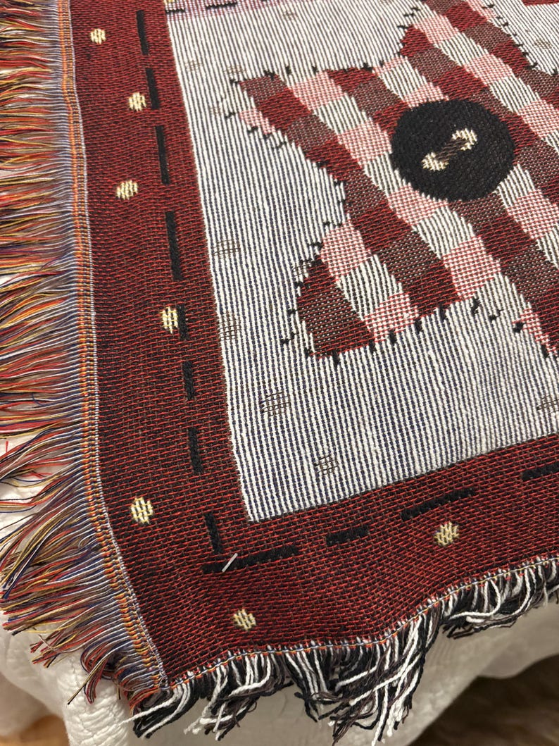 May include: A woven throw blanket with a geometric pattern in shades of red, white, and black. The blanket features a fringed edge with multiple colors. The design includes stripes, squares, and a large black circle.