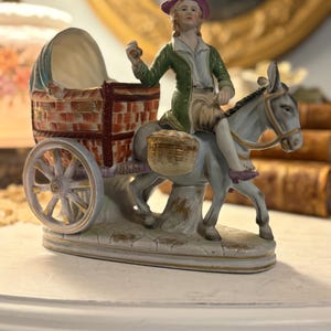 May include: A detailed porcelain figurine depicting a person riding a donkey pulling a cart. The figure wears a green coat and a purple hat. The cart is made of woven material. The figurine is painted in shades of white, brown, green, and gold.