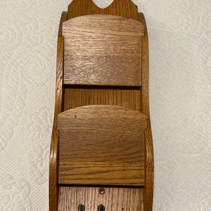 Puede incluir: Organizador de correo de pared de madera con un recorte en forma de corazón en la parte superior. El organizador tiene dos compartimentos y un acabado de veta de madera natural. Diseñado para guardar cartas y otros objetos pequeños.