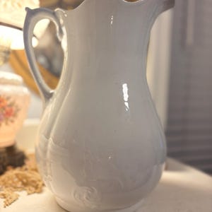 Puede incluir: Una jarra de cerámica blanca con un borde festoneado y un asa curva. La jarra tiene un cuerpo bulboso con sutiles detalles florales en relieve cerca de la base. La jarra es de color blanco suave y parece ser vintage.