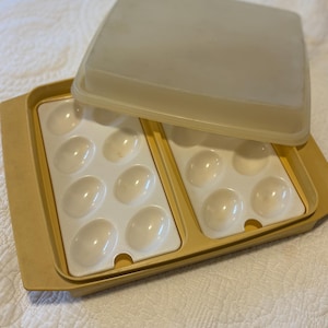 May include: A vintage yellow plastic egg container with a matching lid. The container has two trays, each holding six egg-shaped indentations. The lid is off, revealing the white interior of the egg holders.
