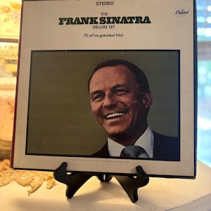 May include: A vintage vinyl record album titled "The Frank Sinatra Deluxe Set" featuring a smiling portrait of Frank Sinatra. The album cover is beige with black and green text, and the Capitol Records logo is visible.