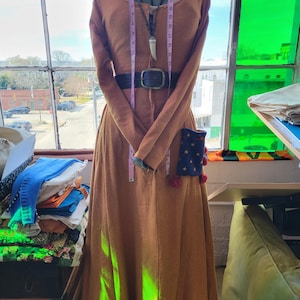 May include: A long, rust-colored dress with long sleeves and a black belt is displayed on a mannequin. The dress has a scoop neckline with blue trim and a small pocket with star and pom-pom details. A measuring tape is draped around the mannequin.