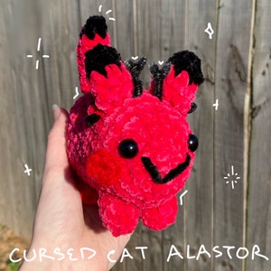 May include: A red and black crocheted cat plush toy with a mischievous expression. The toy has large black eyes and a black mouth with a small smile. The toy is held in a person's hand.