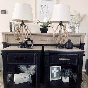 May include: Two black nightstands with a light wood top, each with a drawer and a lower shelf. Gold-toned lamps with white shades sit on top, along with black vases. A framed print with the words "get cozy" hangs in the background.