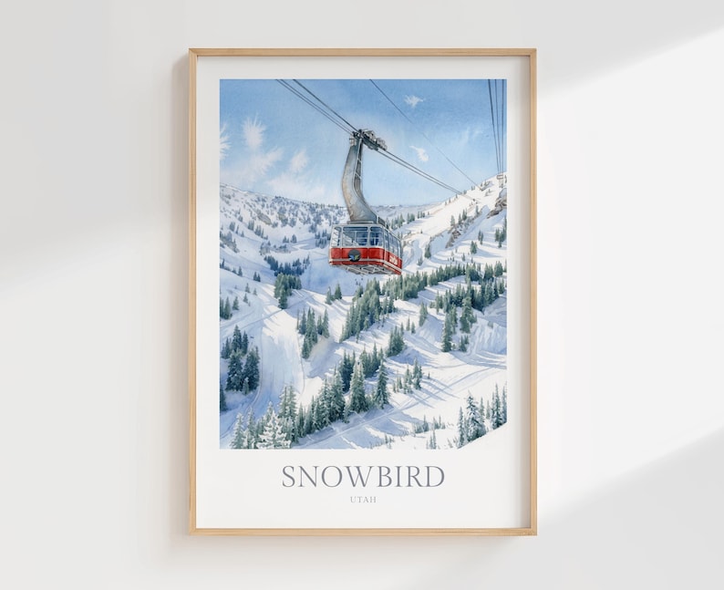 May include: A framed art print featuring a watercolor illustration of a red aerial tram car suspended by cables over a snow-covered mountain landscape with evergreen trees. The text SNOWBIRD UTAH is printed in a grey serif font at the bottom.