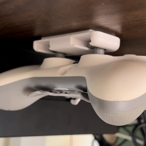 May include: A white video game controller is mounted to a white plastic holder that is attached to the underside of a wooden surface. The controller is facing downwards.
