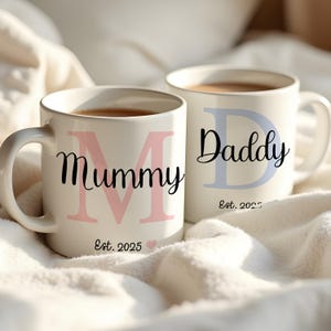 Op de afbeelding: Twee witte keramische mokken met zwarte handvatten. De mok links heeft een roze "M" en de tekst "Mummy Est. 2025". De mok rechts heeft een blauwe "D" en de tekst "Daddy Est. 2025".