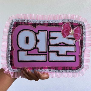 May include: Pink and black plaid sign with white letters that say "이 쪽" surrounded by a pink ruffled trim and a pink bow with a rhinestone in the center.