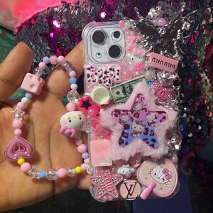 May include: A clear phone case decorated with pink, white, and blue embellishments. The case features a fuzzy pink star, a Hello Kitty charm, a small teacup, a dollar bill, and a Louis Vuitton logo. The case is attached to a pink and blue beaded bracelet.