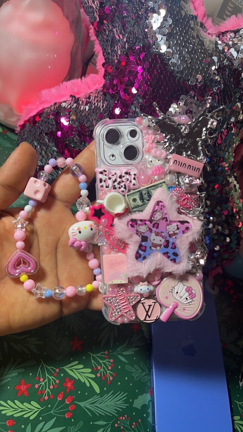 May include: A clear phone case decorated with pink, white, and blue embellishments. The case features a fuzzy pink star, a Hello Kitty charm, a small teacup, a dollar bill, and a Louis Vuitton logo. The case is attached to a pink and blue beaded bracelet.