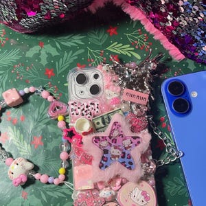 May include: A clear phone case decorated with pink, white, and blue Hello Kitty characters, rhinestones, and a fluffy pink star. The case has a chain attached to it and is sitting on a green background with a pink and silver sequined fabric in the background.