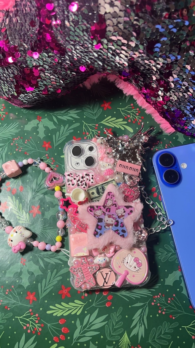 May include: A clear phone case decorated with pink, white, and blue Hello Kitty characters, rhinestones, and a fluffy pink star. The case has a chain attached to it and is sitting on a green background with a pink and silver sequined fabric in the background.