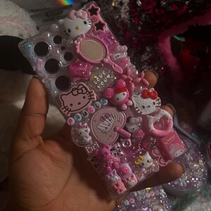 Pu&ograve; includere: Una custodia per telefono rosa e bianca a tema Hello Kitty con uno sfondo trasparente e vari abbellimenti, tra cui ciondoli, strass e glitter. La custodia ha un giocattolo pop-it fidget, uno specchio piccolo e un ciondolo Hello Kitty con il nome "Pamela" scritto su di esso.