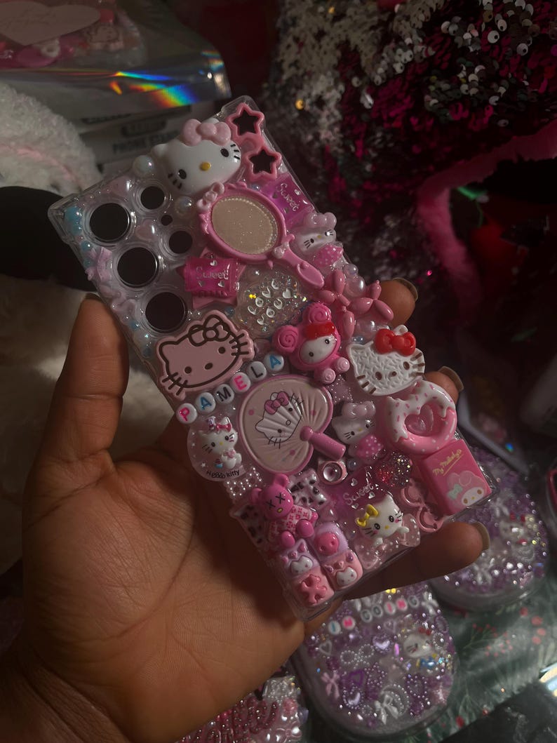 Pu&ograve; includere: Una custodia per telefono rosa e bianca a tema Hello Kitty con uno sfondo trasparente e vari abbellimenti, tra cui ciondoli, strass e glitter. La custodia ha un giocattolo pop-it fidget, uno specchio piccolo e un ciondolo Hello Kitty con il nome "Pamela" scritto su di esso.