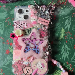 May include: A clear phone case decorated with pink, white, and blue embellishments. The case features a Hello Kitty design, a star with a leopard print, and a charm with the Louis Vuitton logo. The case is attached to a pink Hello Kitty charm and a beaded chain.