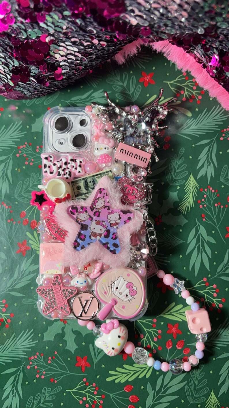May include: A clear phone case decorated with pink, white, and blue embellishments. The case features a Hello Kitty design, a star with a leopard print, and a charm with the Louis Vuitton logo. The case is attached to a pink Hello Kitty charm and a beaded chain.