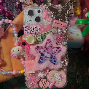 May include: A pink and white phone case decorated with Hello Kitty characters, rhinestones, a pink star, a pink fluffy pom-pom, and a pink and white fan. The case also features a dollar bill, a pink teacup, and a silver butterfly.