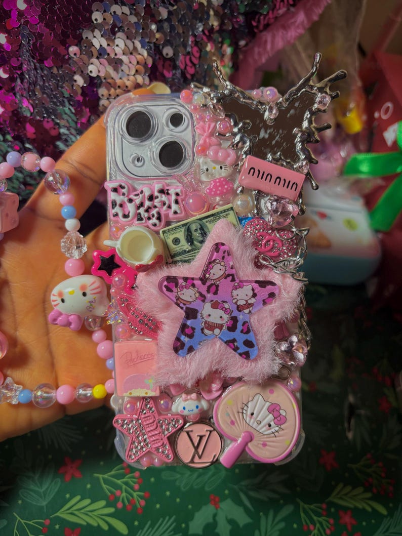 May include: A pink and white phone case decorated with Hello Kitty characters, rhinestones, a pink star, a pink fluffy pom-pom, and a pink and white fan. The case also features a dollar bill, a pink teacup, and a silver butterfly.