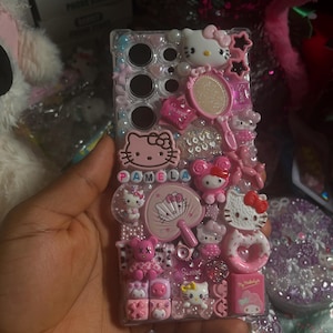 Pu&ograve; includere: Una custodia per telefono rosa Hello Kitty con un retro trasparente e una variet&agrave; di ciondoli Hello Kitty, tra cui uno specchio, un ventaglio e un cuore. La custodia ha anche il nome "Pamela" scritto in lettere rosa.