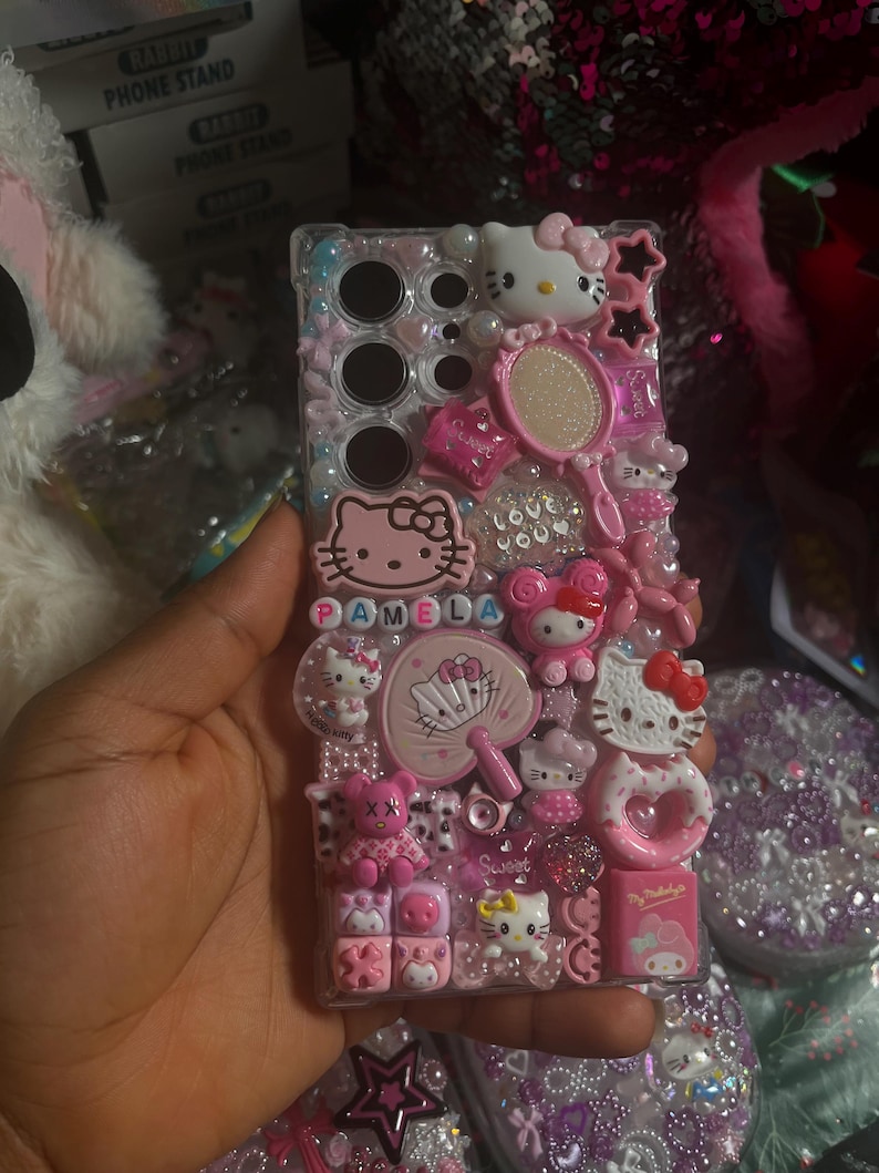 Pu&ograve; includere: Una custodia per telefono rosa Hello Kitty con un retro trasparente e una variet&agrave; di ciondoli Hello Kitty, tra cui uno specchio, un ventaglio e un cuore. La custodia ha anche il nome "Pamela" scritto in lettere rosa.