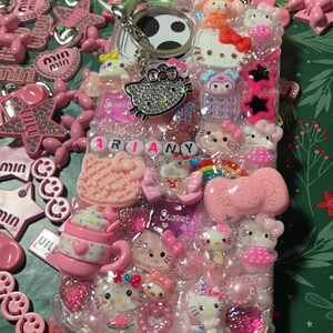 Pu&ograve; includere: Una custodia per telefono rosa e trasparente decorata con personaggi di Hello Kitty, strass e altri ciondoli. La custodia ha un anello porta-dito bianco e nero e il nome "ARIANY" scritto in lettere rosa.