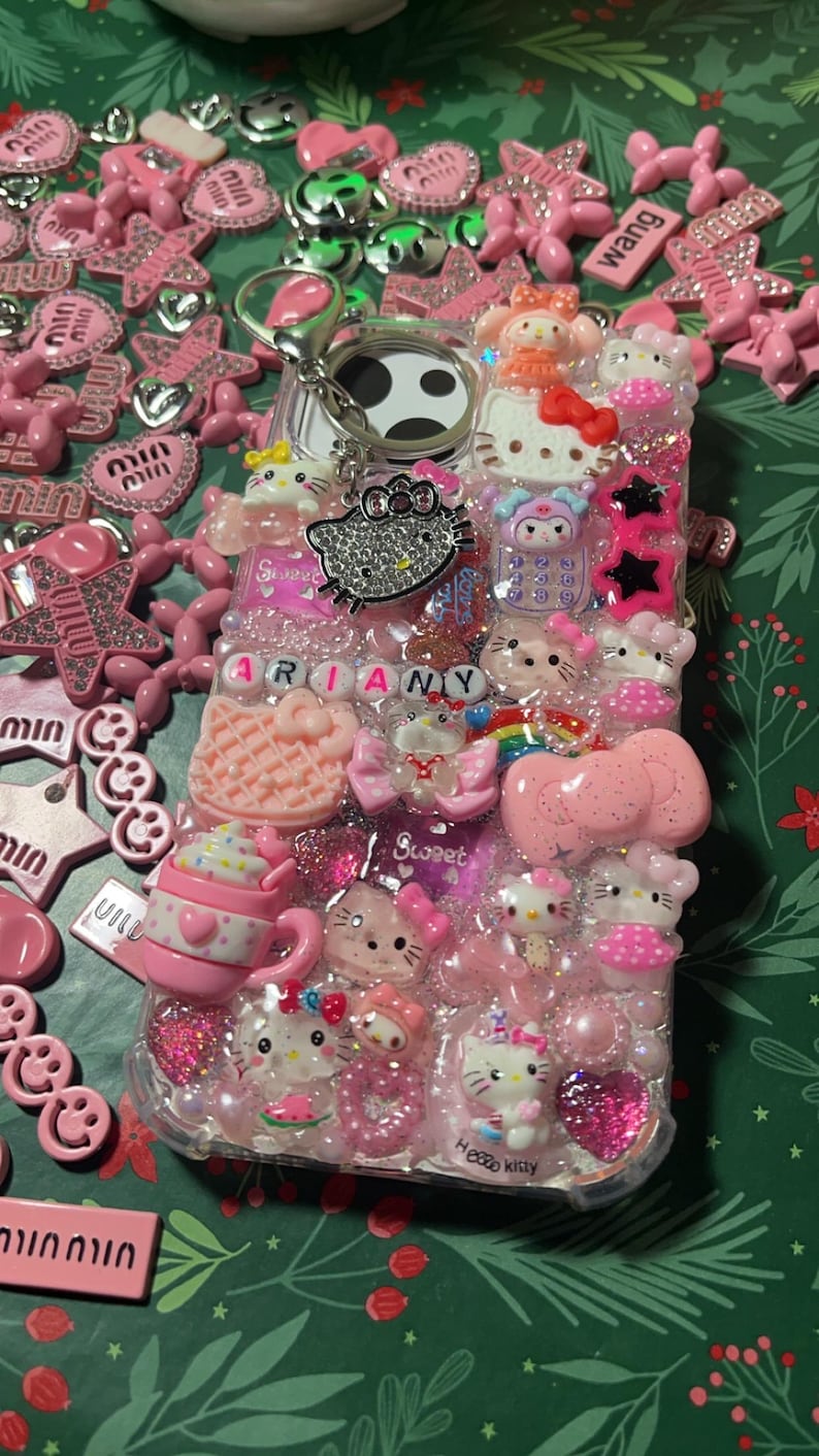 Pu&ograve; includere: Una custodia per telefono rosa e trasparente decorata con personaggi di Hello Kitty, strass e altri ciondoli. La custodia ha un anello porta-dito bianco e nero e il nome "ARIANY" scritto in lettere rosa.