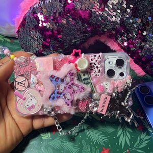 May include: A clear phone case decorated with pink, white, and blue embellishments. The case features a Hello Kitty character, a star-shaped charm, and a chain strap. The case is covered in glitter and rhinestones.