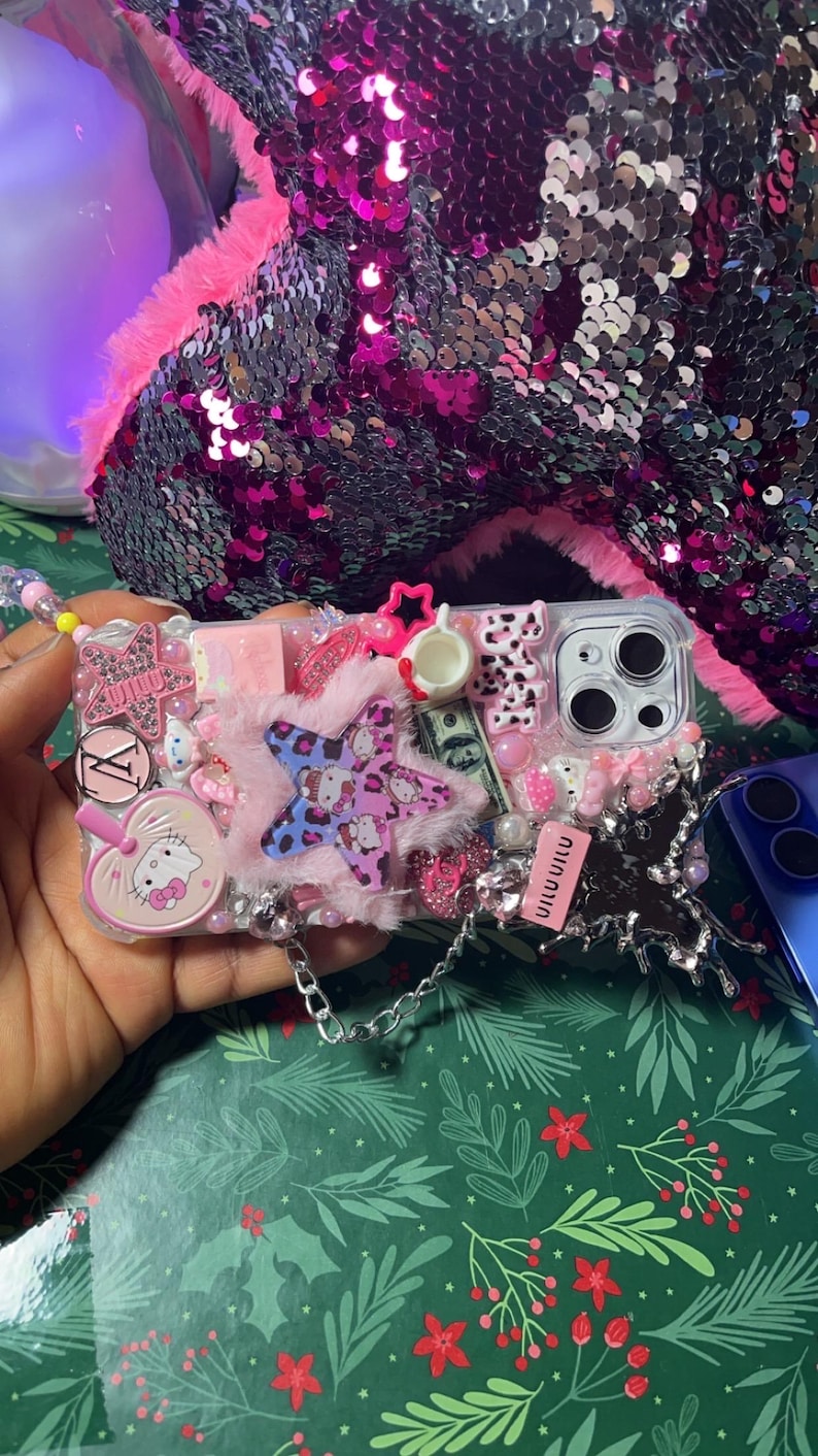 May include: A clear phone case decorated with pink, white, and blue embellishments. The case features a Hello Kitty character, a star-shaped charm, and a chain strap. The case is covered in glitter and rhinestones.