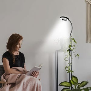 May include: A black and silver reading lamp with a circular light head illuminates a plant. The lamp is positioned next to a woman reading a book while wrapped in a pink blanket. The background is a white wall.