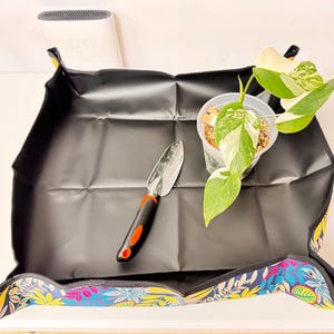 May include: A black gardening mat with a colourful floral border, a small trowel, and a potted plant with green and white leaves. The mat is designed to contain soil and water during repotting or other gardening tasks.
