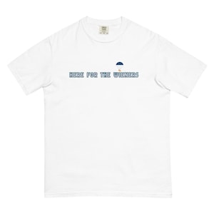 Può includere: T-shirt bianca con la scritta blu "HERE FOR THE WIENERS" e una piccola illustrazione di un paracadute. Un capo d'abbigliamento casual.