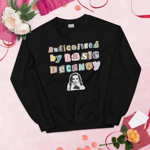 Puede incluir: Sudadera negra con el texto "Radicalized by Basic Decency" en letras coloridas de estilo recortado. Debajo del texto hay una ilustración en blanco y negro de una mujer cubriéndose la boca. La sudadera es de cuello redondo.