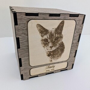 May include: A wooden pet memorial box with a laser-etched portrait of a cat. The box is grey with black accents and features the cat's name, "Bailey," and the years "2006-2025" engraved on the front. The box is cube-shaped.