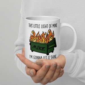 Op de afbeelding: Witte keramische mok met een ontwerp van een brandende groene vuilcontainer met de naam "Mike". De mok heeft de tekst "THIS LITTLE LIGHT OF MINE I'M GONNA LET IT SHINE" in het zwart. Een humoristisch cadeau-idee.