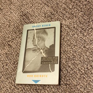 Peut inclure: Un livre de poche intitulé "Don Quichotte : Un roman" de Kathy Acker. La couverture présente une image en noir et blanc d'une personne avec un tatouage, sur fond bleu clair. Inclut une citation du New York Times Book Review.