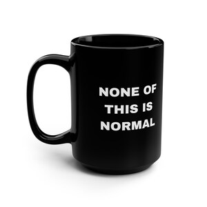 May include: Black ceramic mug with a large handle. The mug features the white text "NONE OF THIS IS NORMAL" in a bold, sans-serif font. The mug is a standard size for tea or coffee.