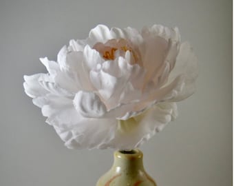 May include: A single white peony with a light pink center in a small, hand-thrown ceramic vase with a light brown glaze and orange stripes. "Super light pink | Open" is written below the vase.