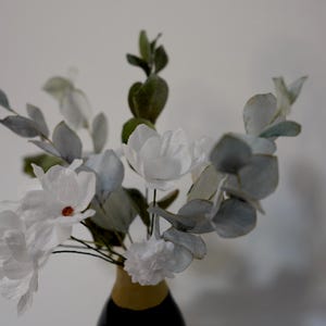 May include: A bouquet of white paper flowers with green and grey eucalyptus leaves in a brown vase.