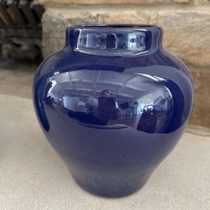 May include: A glossy, navy blue ceramic vase with a rounded body and a narrow neck. The vase reflects the surrounding environment, showcasing a smooth, polished surface. The vase is a decorative item.
