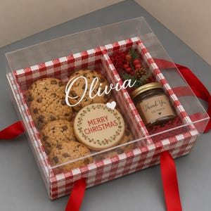 May include: A clear gift box with a red and white gingham pattern containing chocolate chip cookies, a round cookie with the text MERRY CHRISTMAS, and a small glass jar labeled Thank You SO MUCH. The name Olivia is printed on the lid.