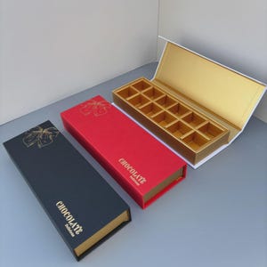 Puede incluir: Tres cajas rectangulares de bombones en negro, rojo y blanco. Las cajas negras y rojas tienen texto dorado que dice "CHOCOLATE Delicious" y un diseño floral dorado. La caja blanca está abierta, revelando un interior dorado con 12 compartimentos.