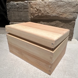 May include: A rectangular light-colored wooden box with a lid. The box features visible finger joints at the corners, showcasing the natural grain and texture of the light tan wood. The lid sits flush on top of the box, creating a clean, simple design.
