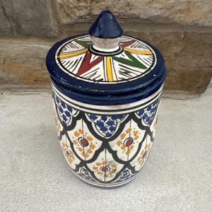 May include: A ceramic jar with a lid, featuring a vibrant, hand-painted design. The jar is primarily white with blue, yellow, red, and green accents. The lid has a geometric pattern, and the jar's body displays a repeating floral motif. The jar is approximately 5 inches tall.